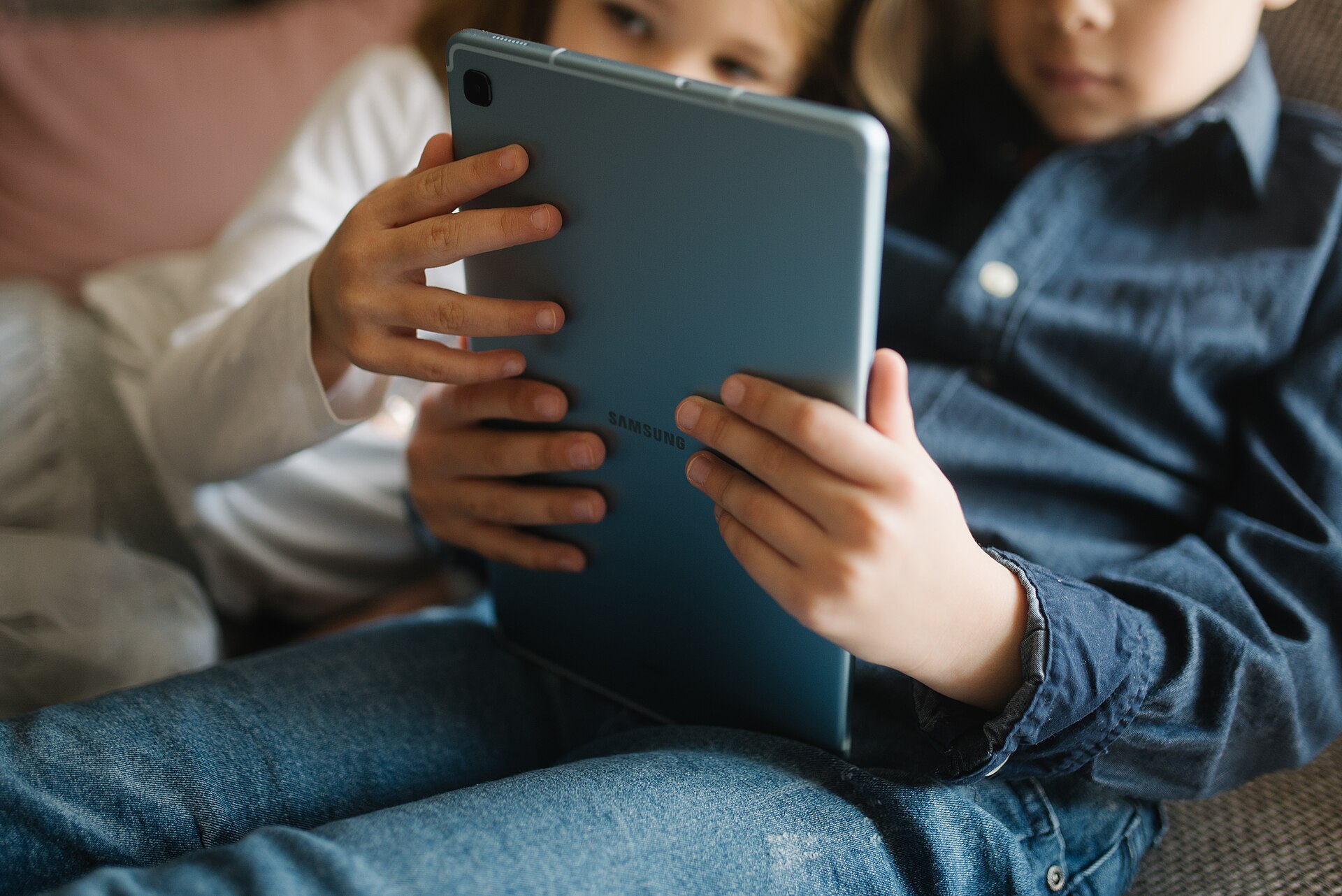 Two children on a living-room couch, looking at a tablet held between them.