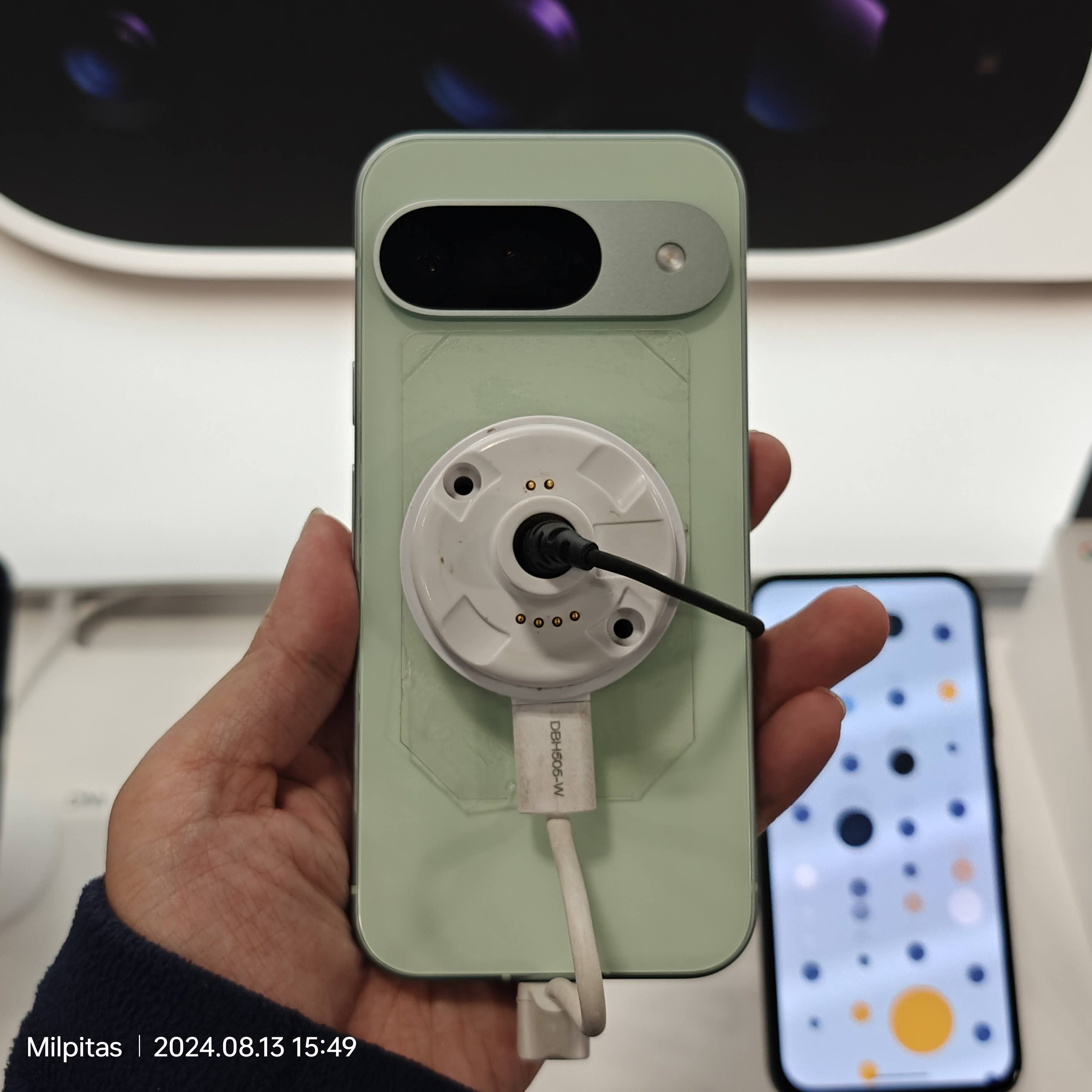 A hand holding the back of a pale green Google Pixel 9 attached to a retail security tether.