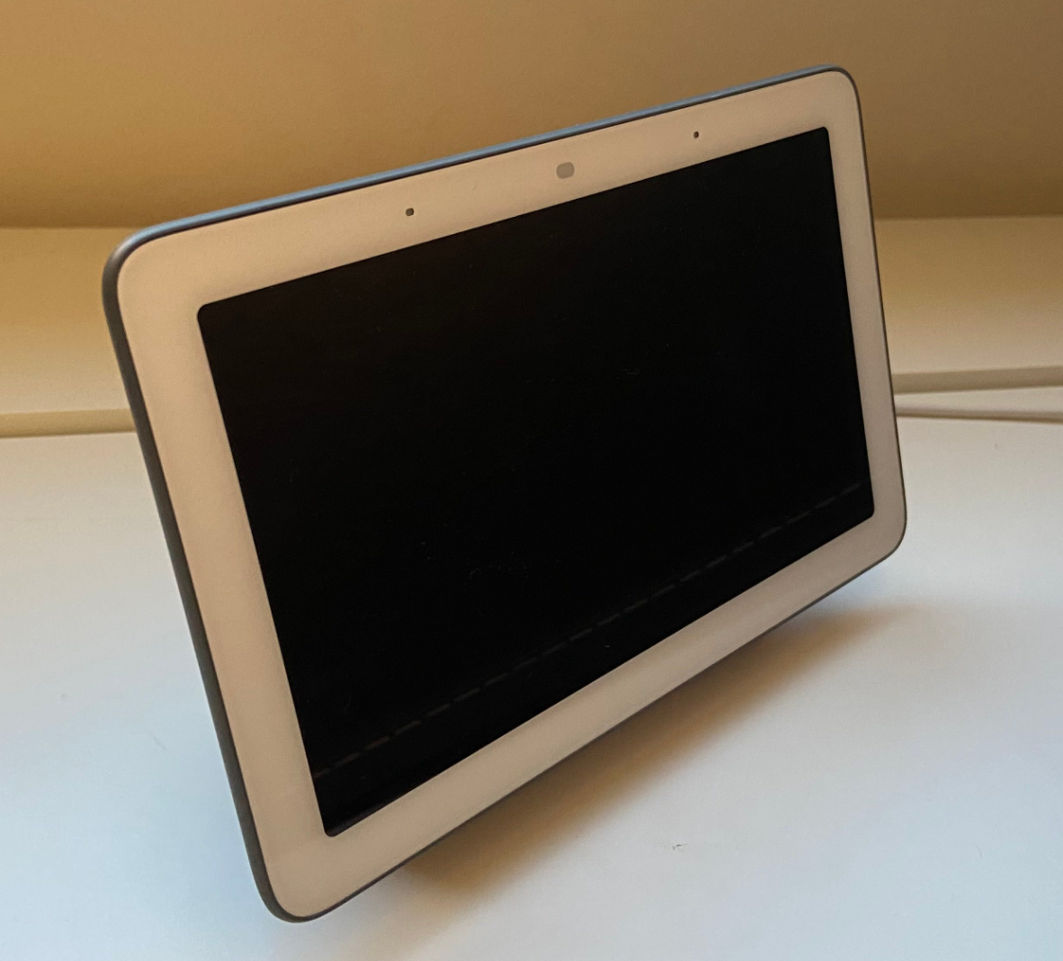 A gray Google Nest Hub smart display with a dark screen, sitting on a desk with its power cord behind it.