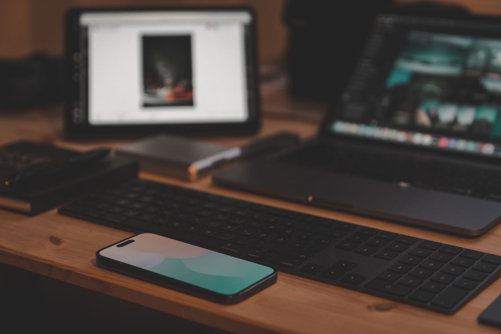 A laptop, tablet and smartphone arranged on a wooden desk beside a black speaker.
