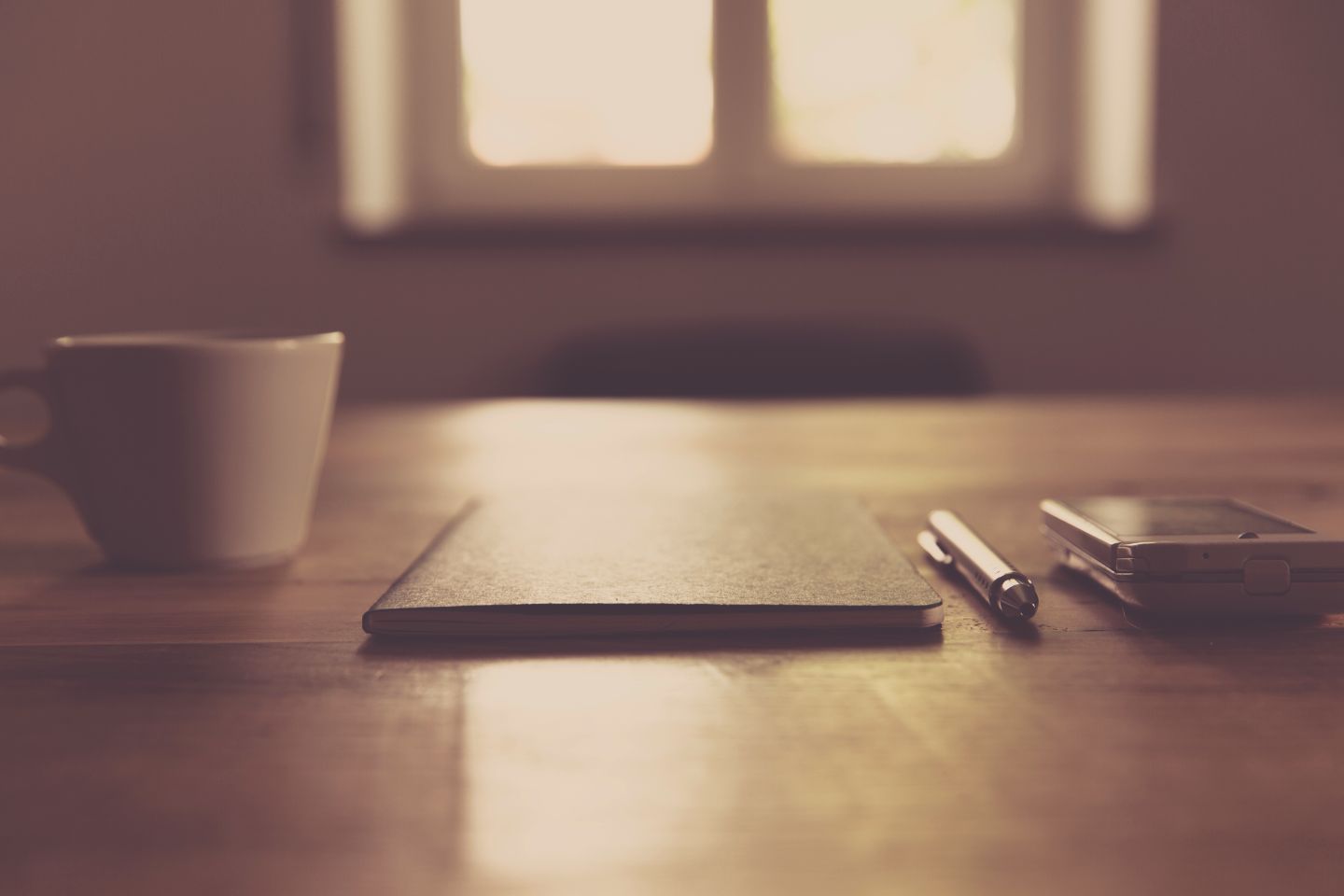 A smartphone beside a closed notebook, pen and white cup on a wooden desk.