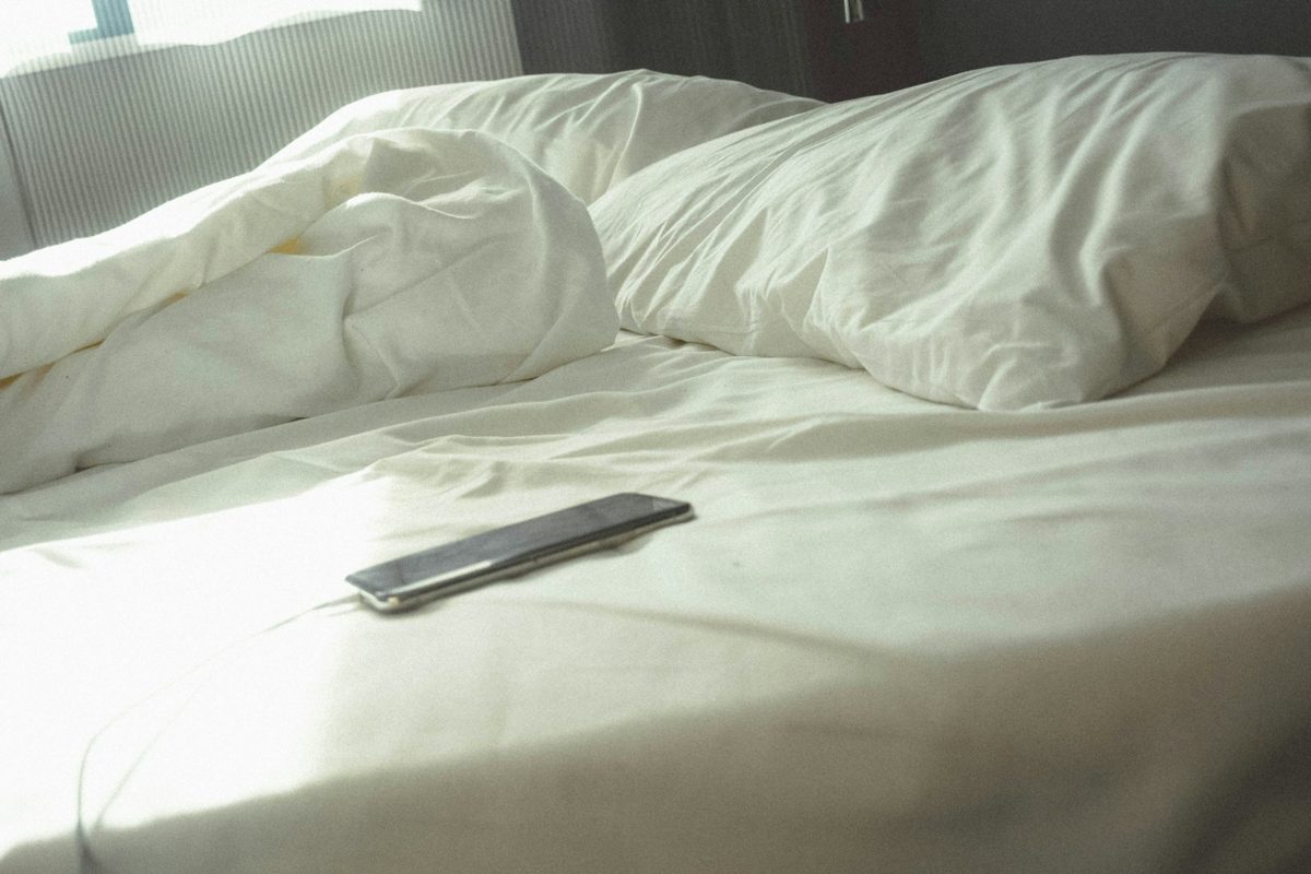 A black smartphone plugged into a cable rests on white bedding beside pillows.