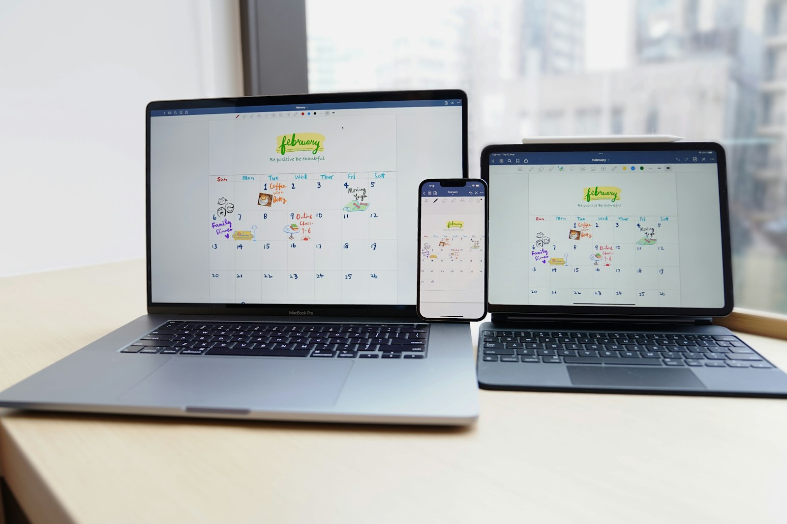 A laptop, phone and tablet displaying the same handwritten February calendar on a wooden desk.