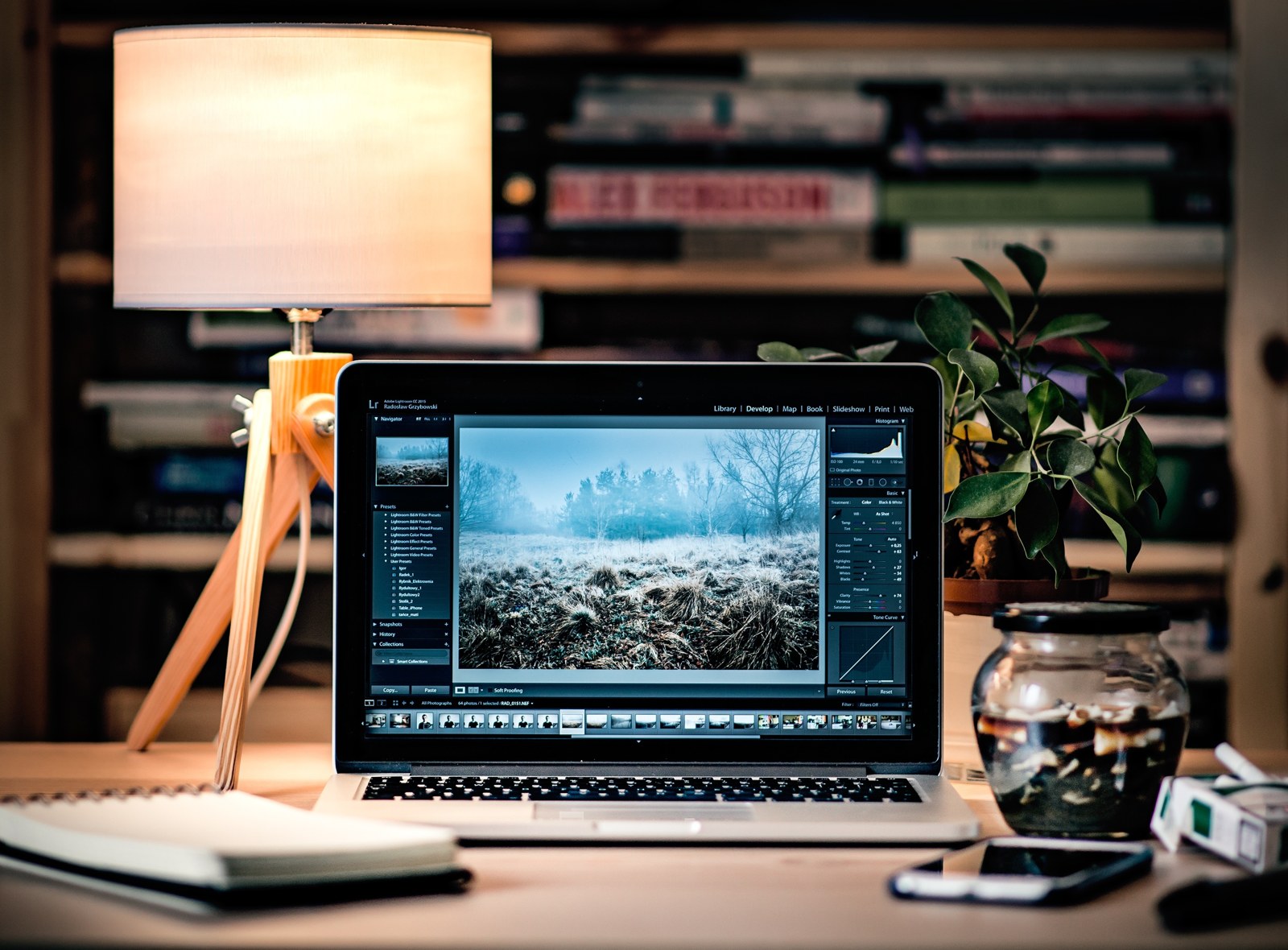 A laptop showing photo-editing software sits beside a lamp and plant on a desk.