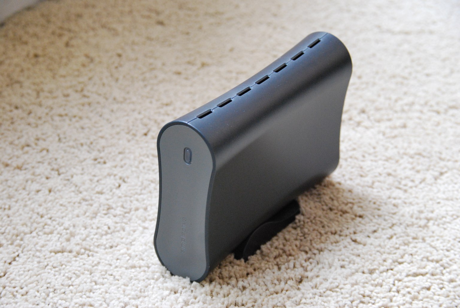 A dark external hard drive stands upright on a cream-colored carpet.