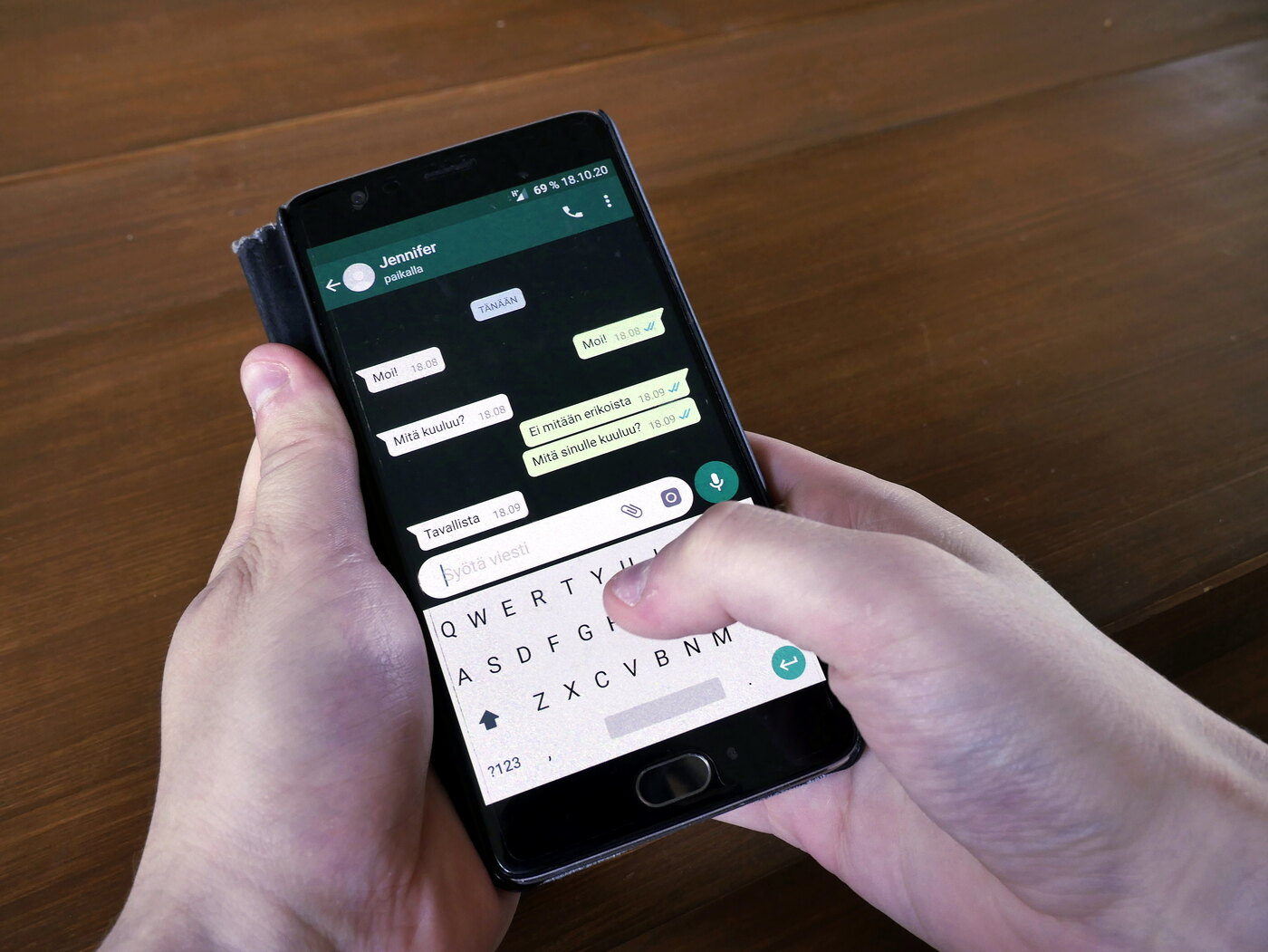 Hands holding a phone with a WhatsApp text conversation above a wooden table, photographed in 2018.