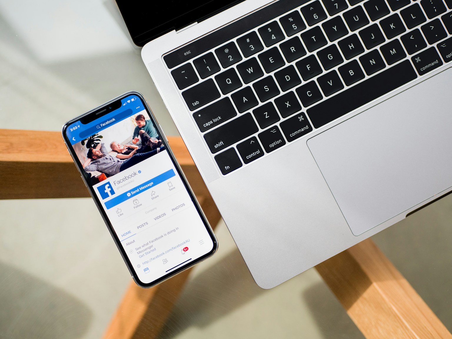 An iPhone displaying a Facebook page beside a silver MacBook on a glass table.