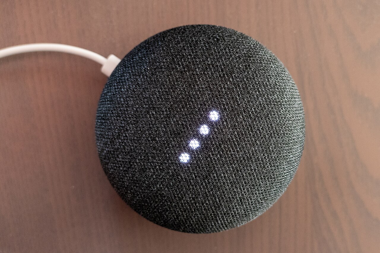A charcoal Google Home Mini speaker with four illuminated dots and a white power cable on a wooden surface.