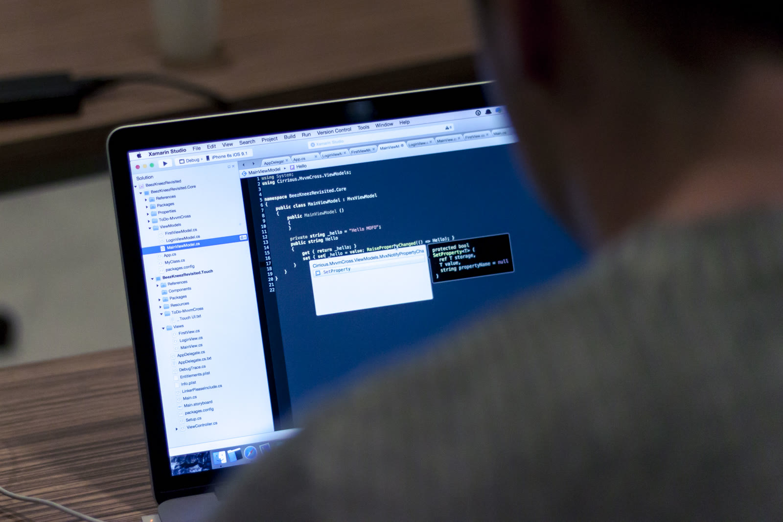 An open laptop on a wooden desk beside a notebook of handwritten notes, with programming code on the screen and a small plant nearby.