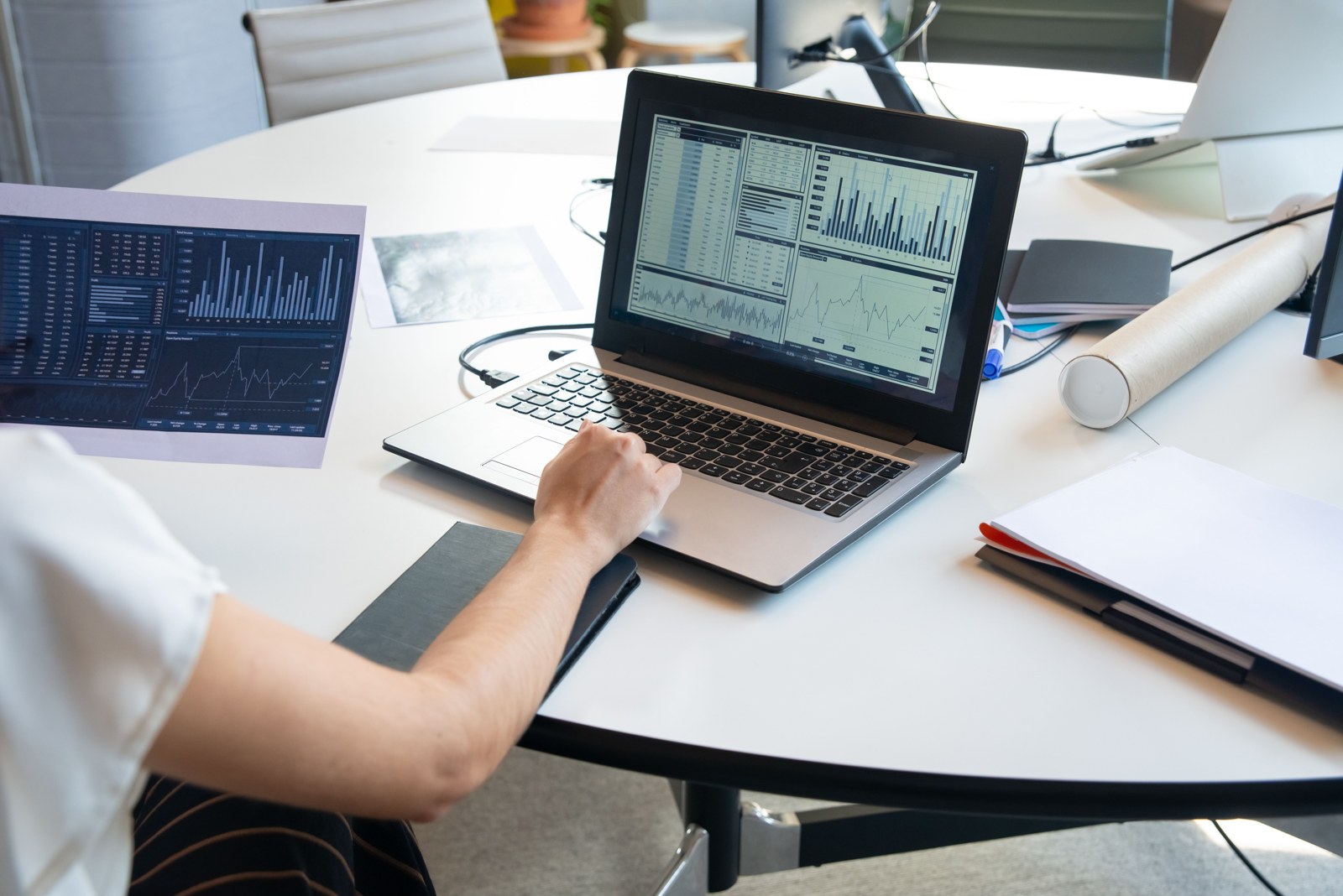 A person uses a laptop displaying charts beside a printed report on a white office table.