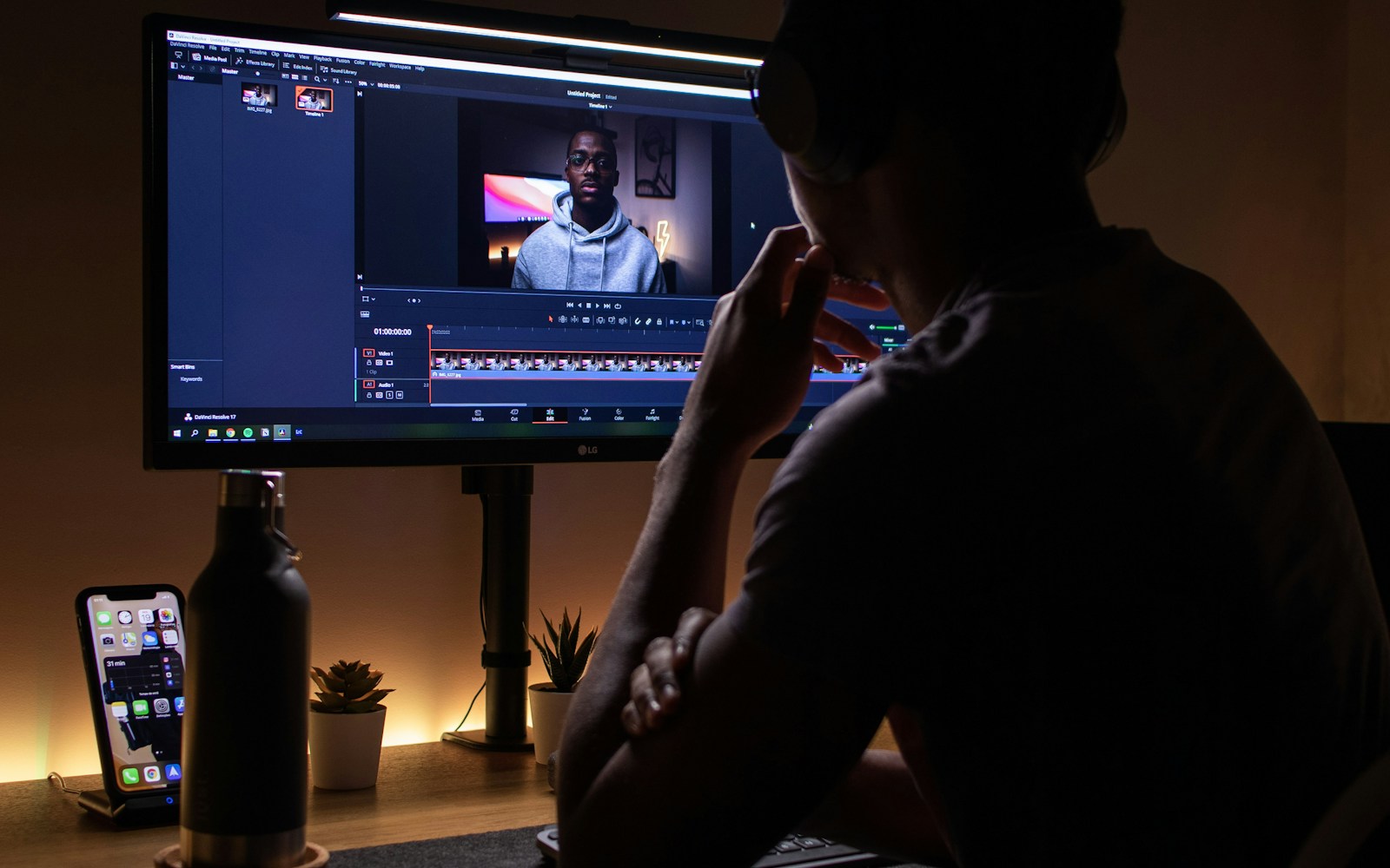 A person wearing headphones sits at a monitor showing a video-editing timeline and a talking-head recording.