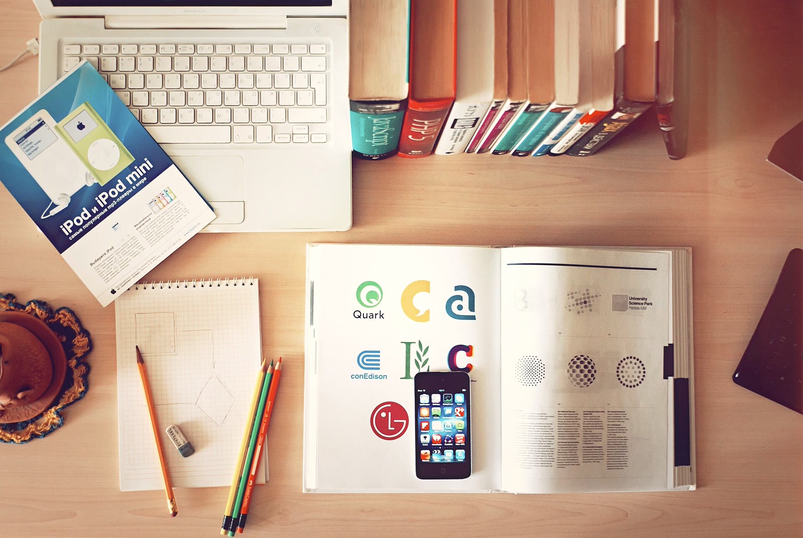 An open laptop on a wooden desk beside stacked books, colored pencils, and a smartphone.