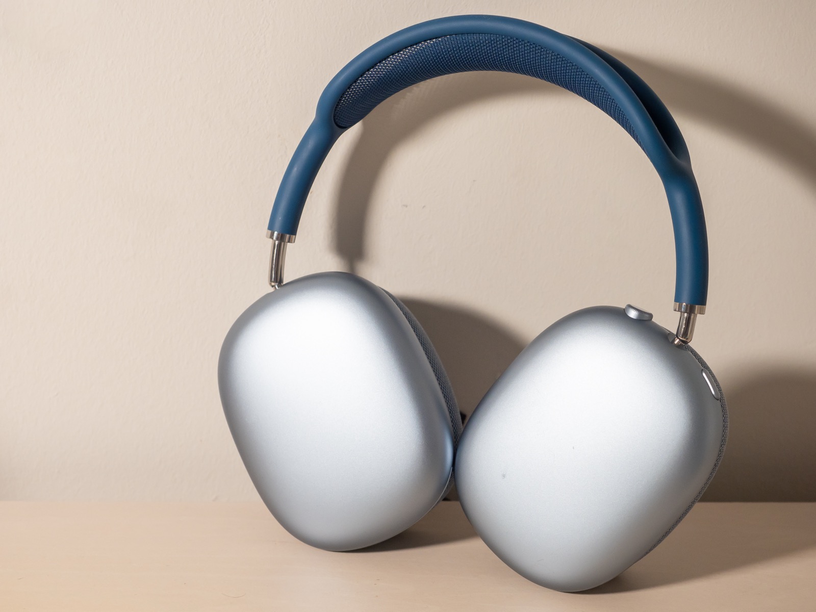 Silver AirPods Max headphones with a blue headband resting upright on a light surface.