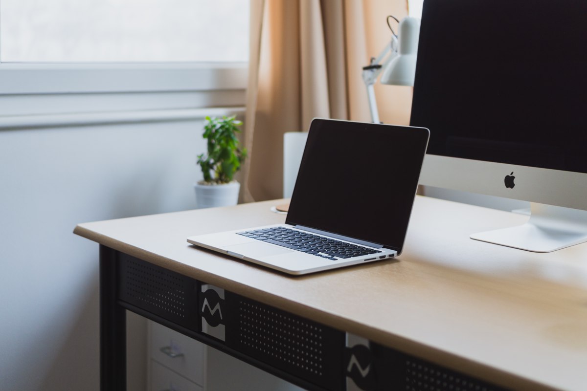 An open silver MacBook on a wooden desk beside an iMac, a lamp and a small plant.
