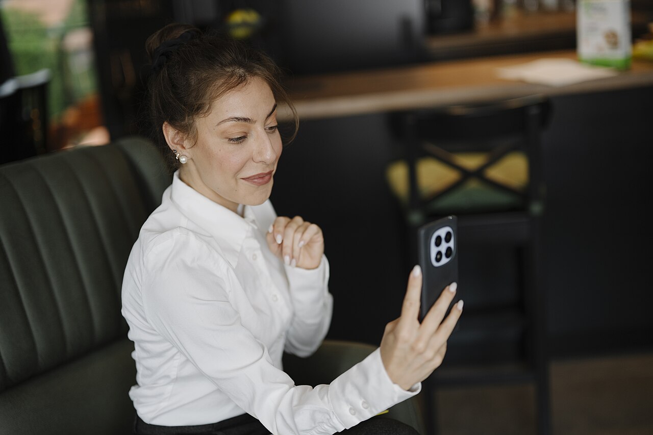 A woman seated in a cafe holding a smartphone in front of her during a video call.