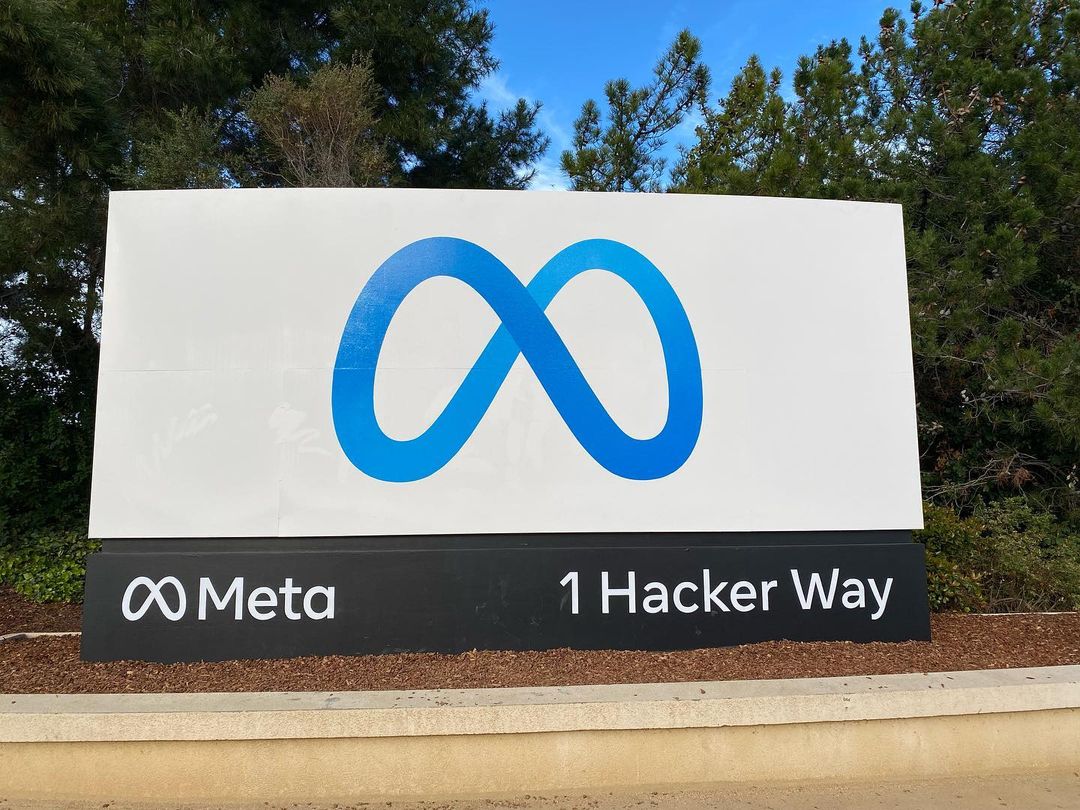 The blue Meta sign at the entrance of Meta's Menlo Park headquarters, with an office building, parked cars, and trees behind it.