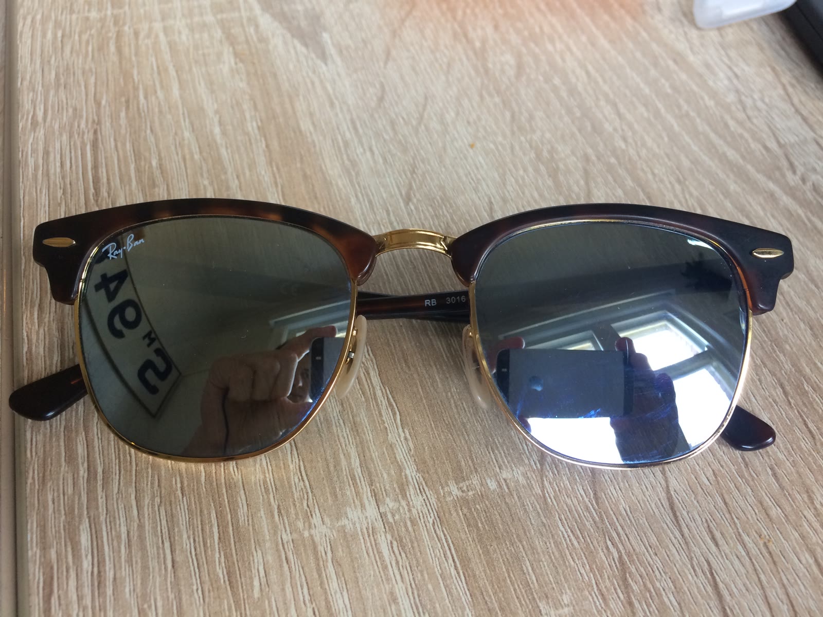 A pair of Ray-Ban Clubmaster sunglasses with a tortoiseshell browline, gold-colored lower rims, and dark lenses, lying on a pale surface.