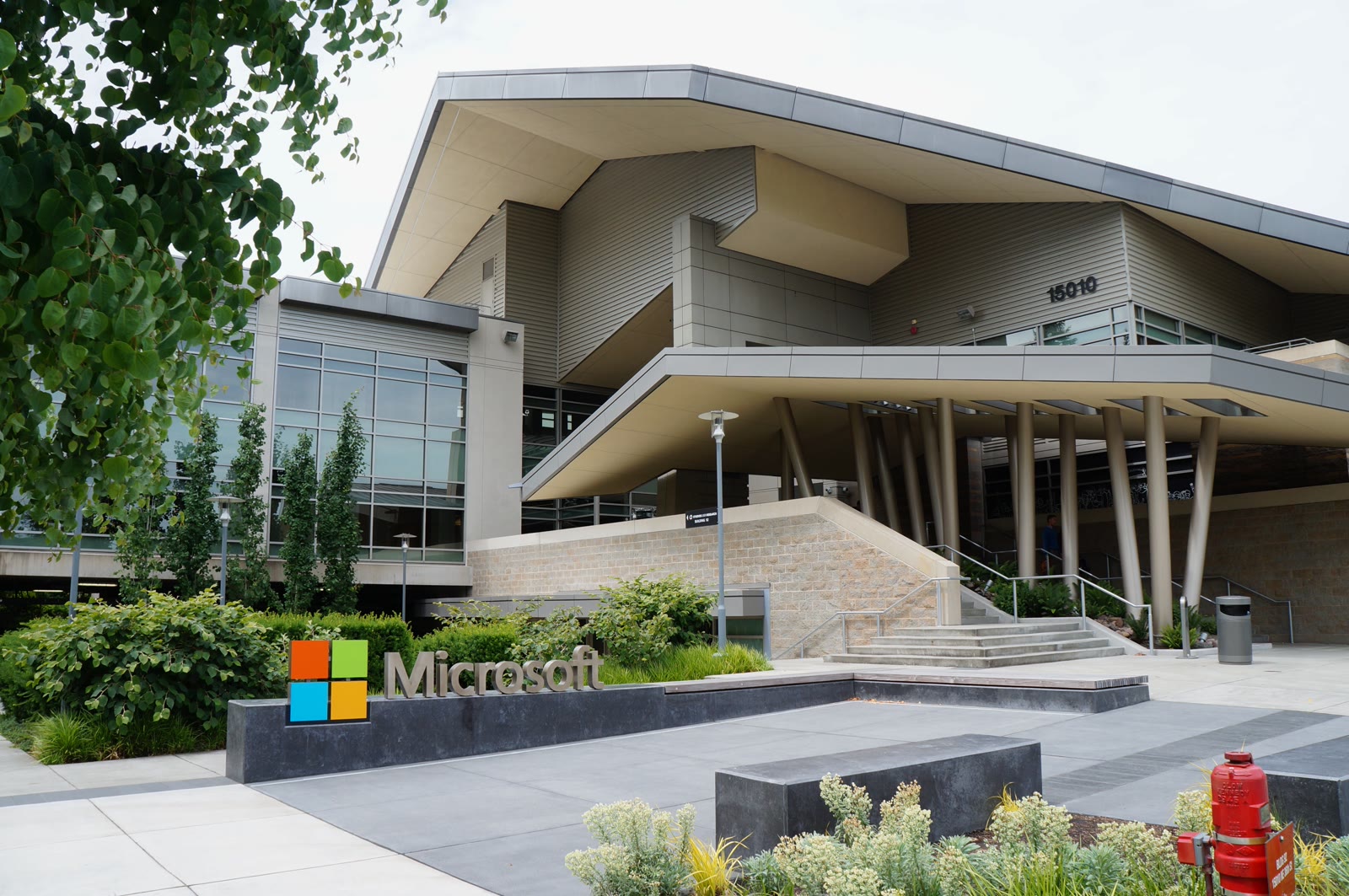 Building 92 on Microsoft's Redmond campus, a curved glass office with the Microsoft logo on the facade, trees along the sidewalk, and a blue sky with light clouds.