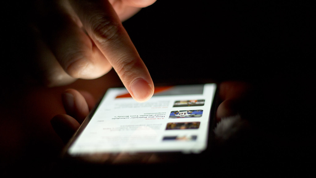 A hand scrolls a list of news stories on a smartphone in a dark room, the screen lighting the fingers.