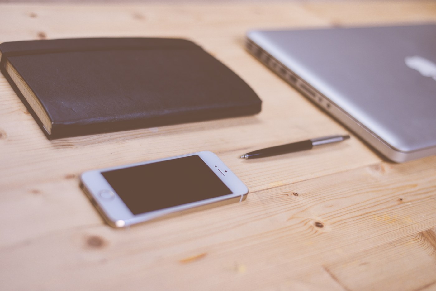 A white smartphone with its screen dark on a wooden desk beside a notebook, pen and closed laptop.
