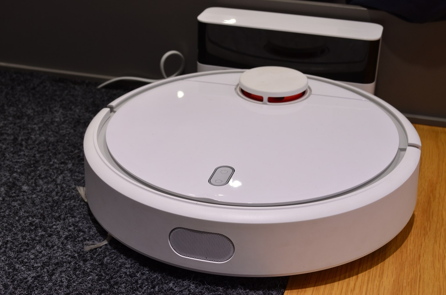 A white Xiaomi Mi Robot Vacuum Cleaner in front of its charging dock, partly on a dark rug beside a wood floor.
