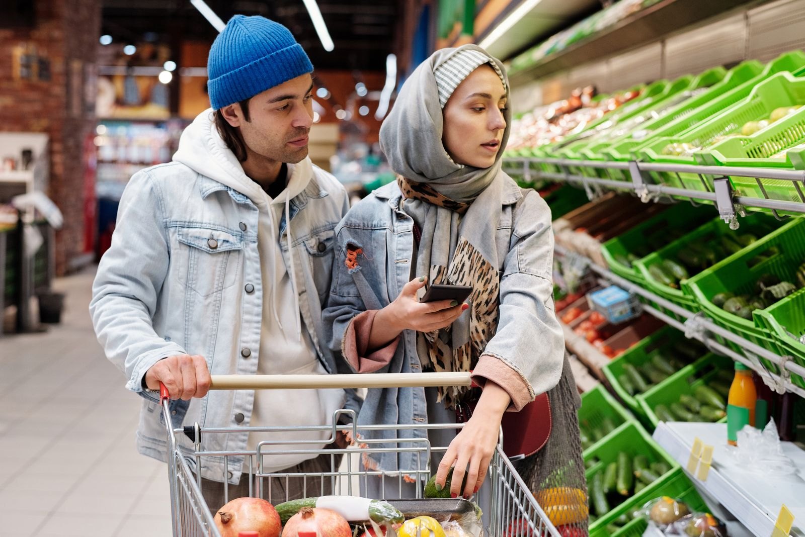 Two shoppers stand beside a grocery cart in a supermarket produce aisle, with one holding a smartphone.