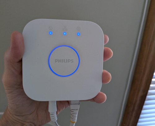 A hand holds a white Philips Hue Bridge with blue indicator lights and connected cables.