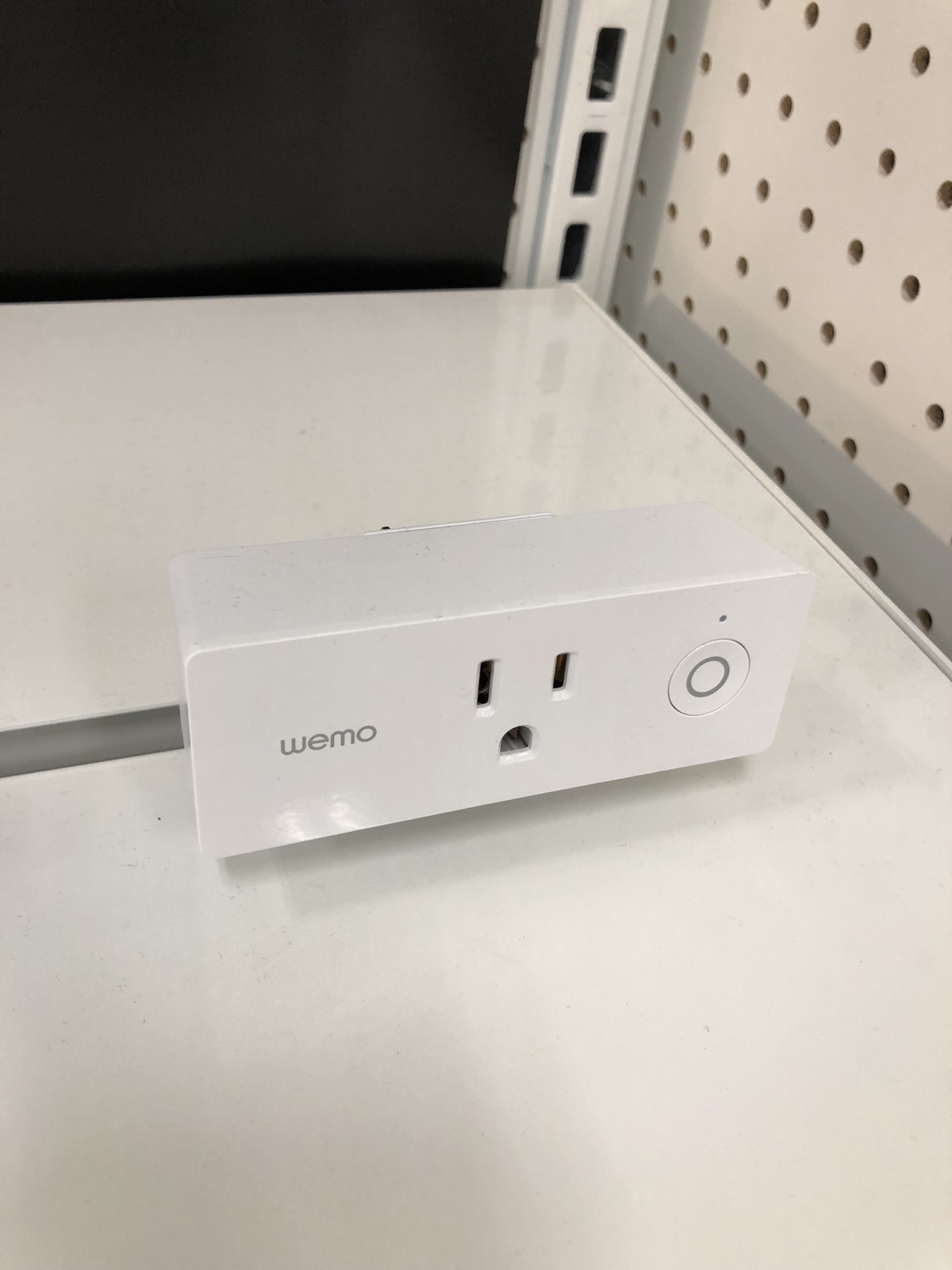 A white Wemo smart plug with a three-slot outlet and round power button resting on a white shelf.
