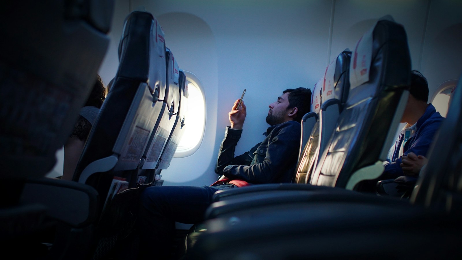 A passenger seated beside an airplane window looks at a smartphone.