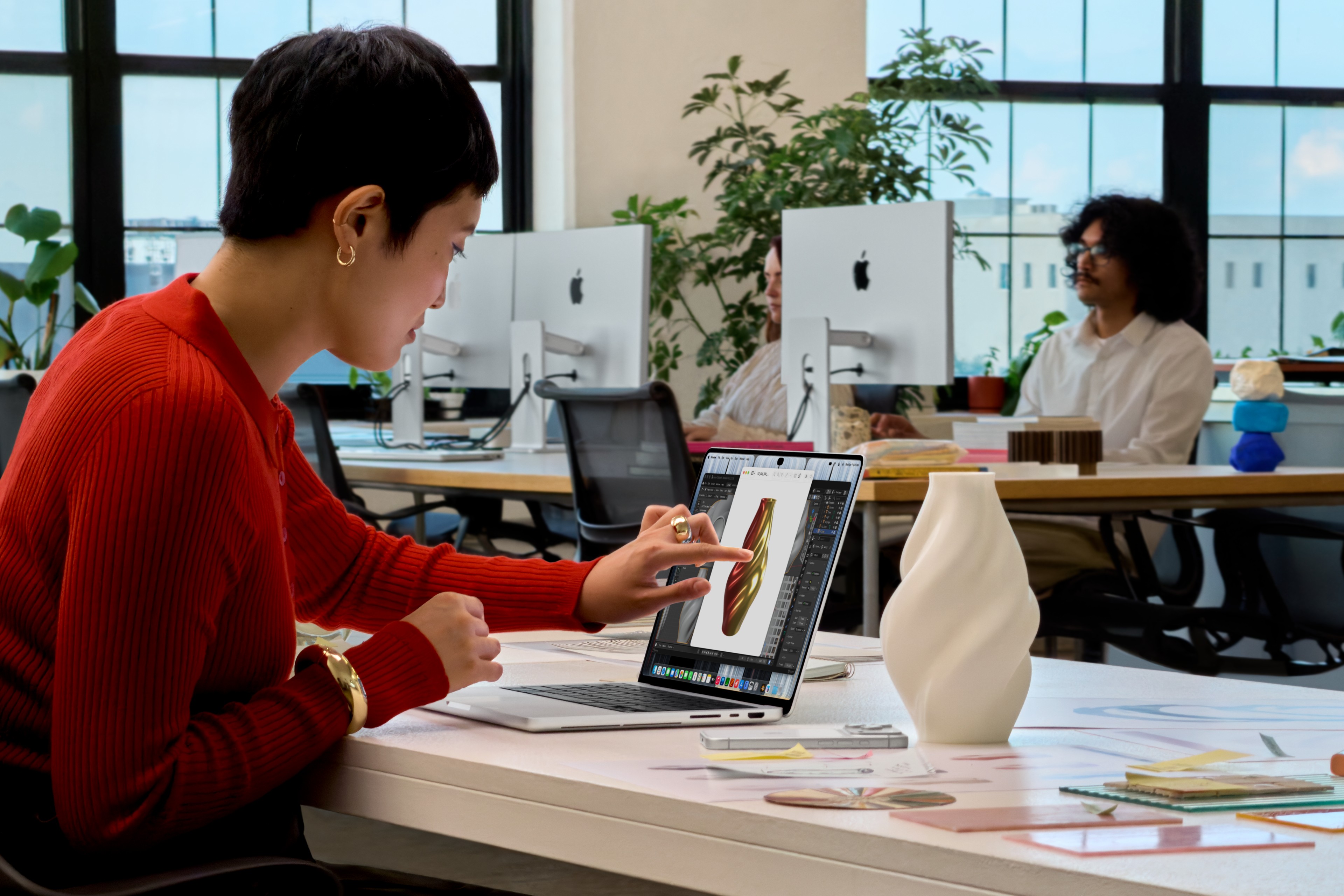 A person in a red top touches a silver MacBook Pro’s screen while working on a vase design at an office desk.