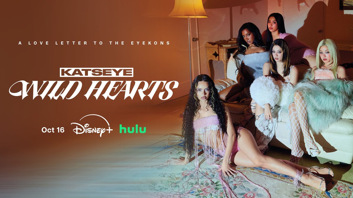 Official KATSEYE: WILD HEARTS key art showing the six group members together in a warmly lit room.