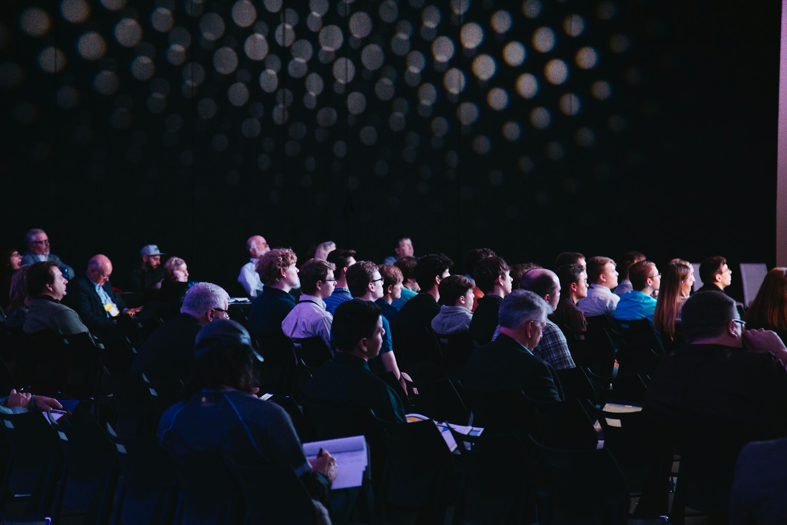 Audience seated in a darkened conference hall facing a bright stage