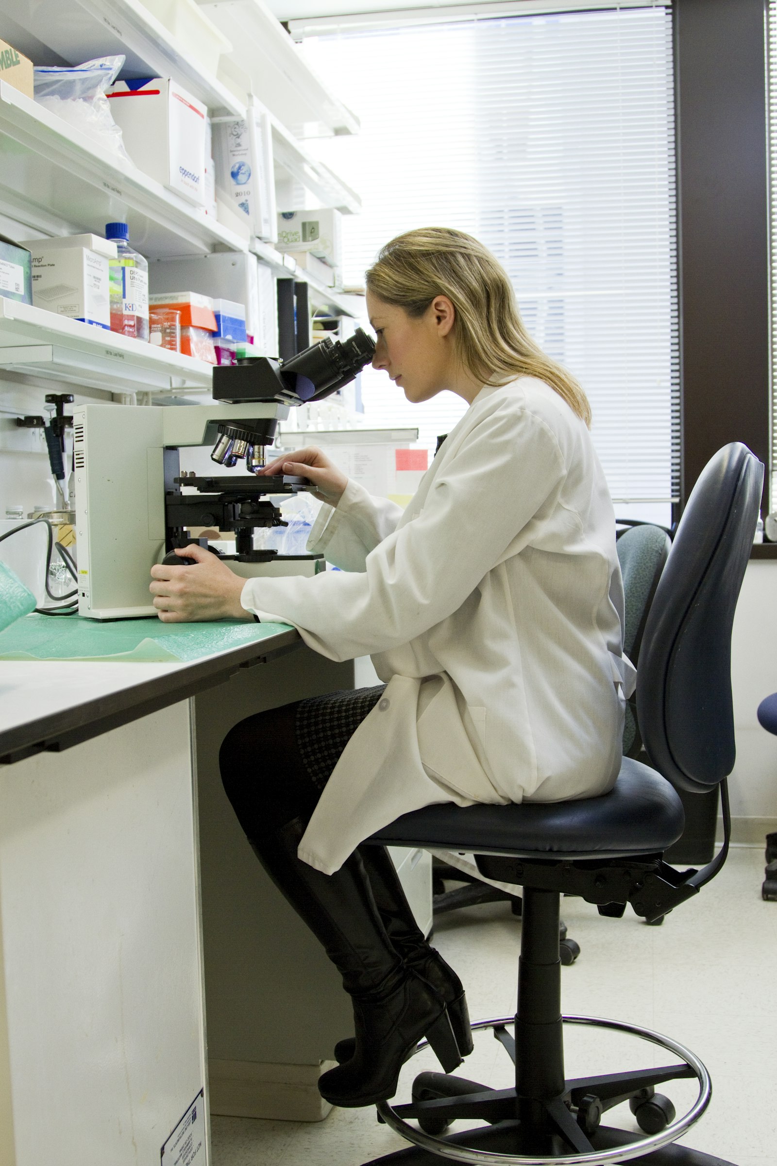 Scientist in a white lab coat looking through a compound microscope on a lab bench