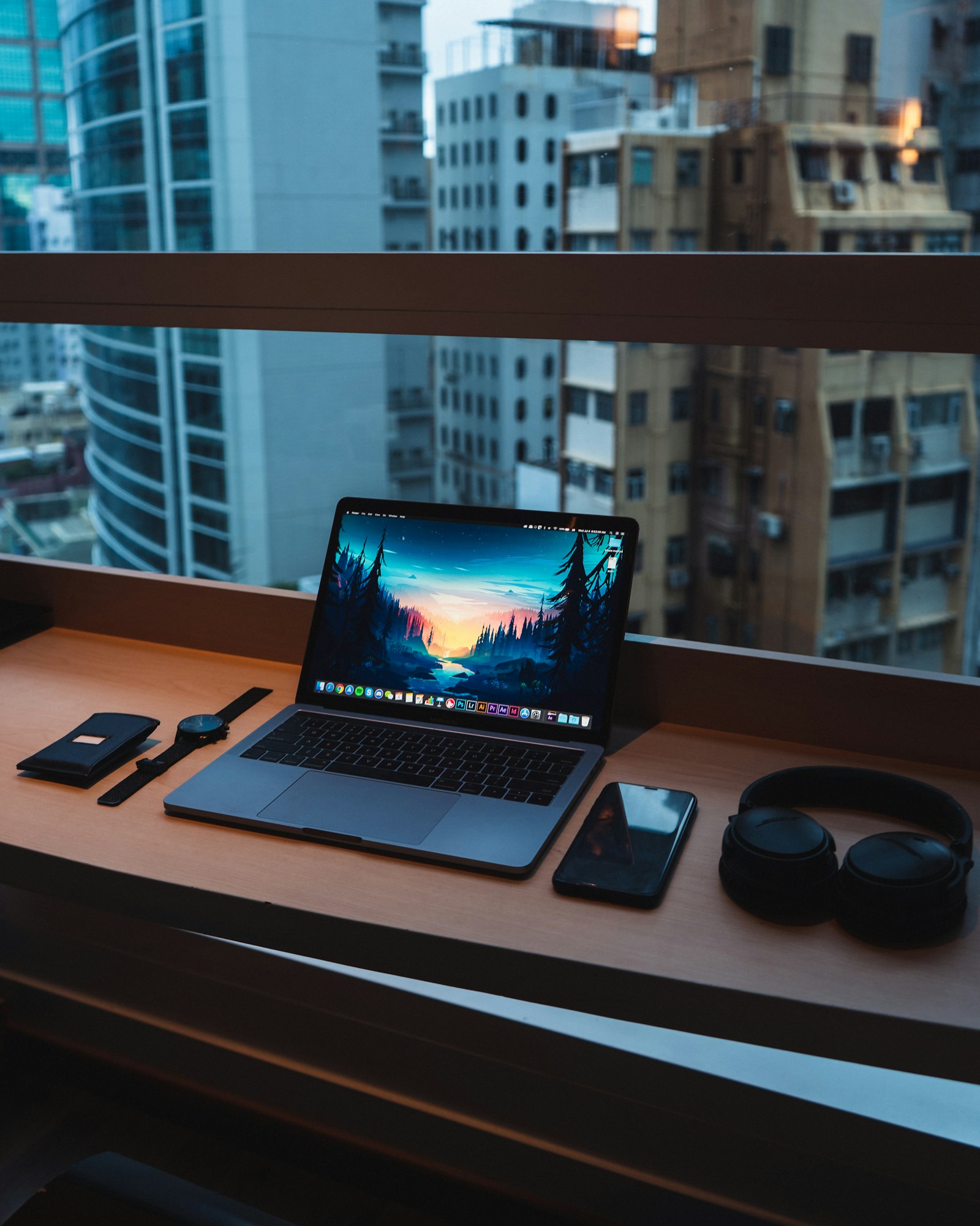 Open MacBook on a desk with headphones and phone by a city window