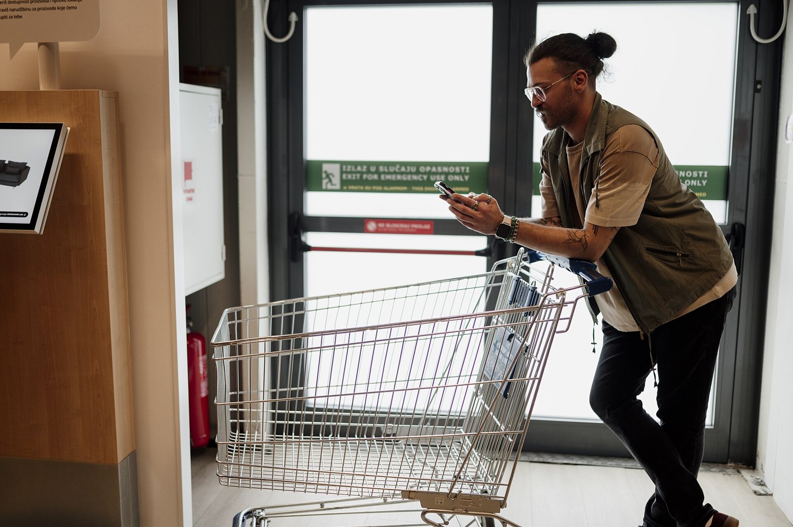 A shopper leans on a shopping cart while checking his phone near a store exit