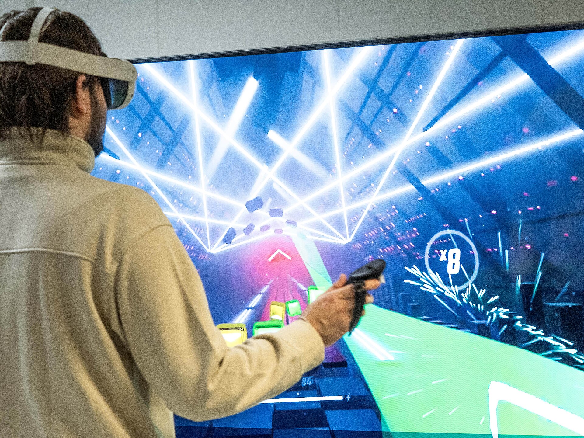 Player wearing a white standalone VR headset swings a controller in a rhythm game shown on a large screen