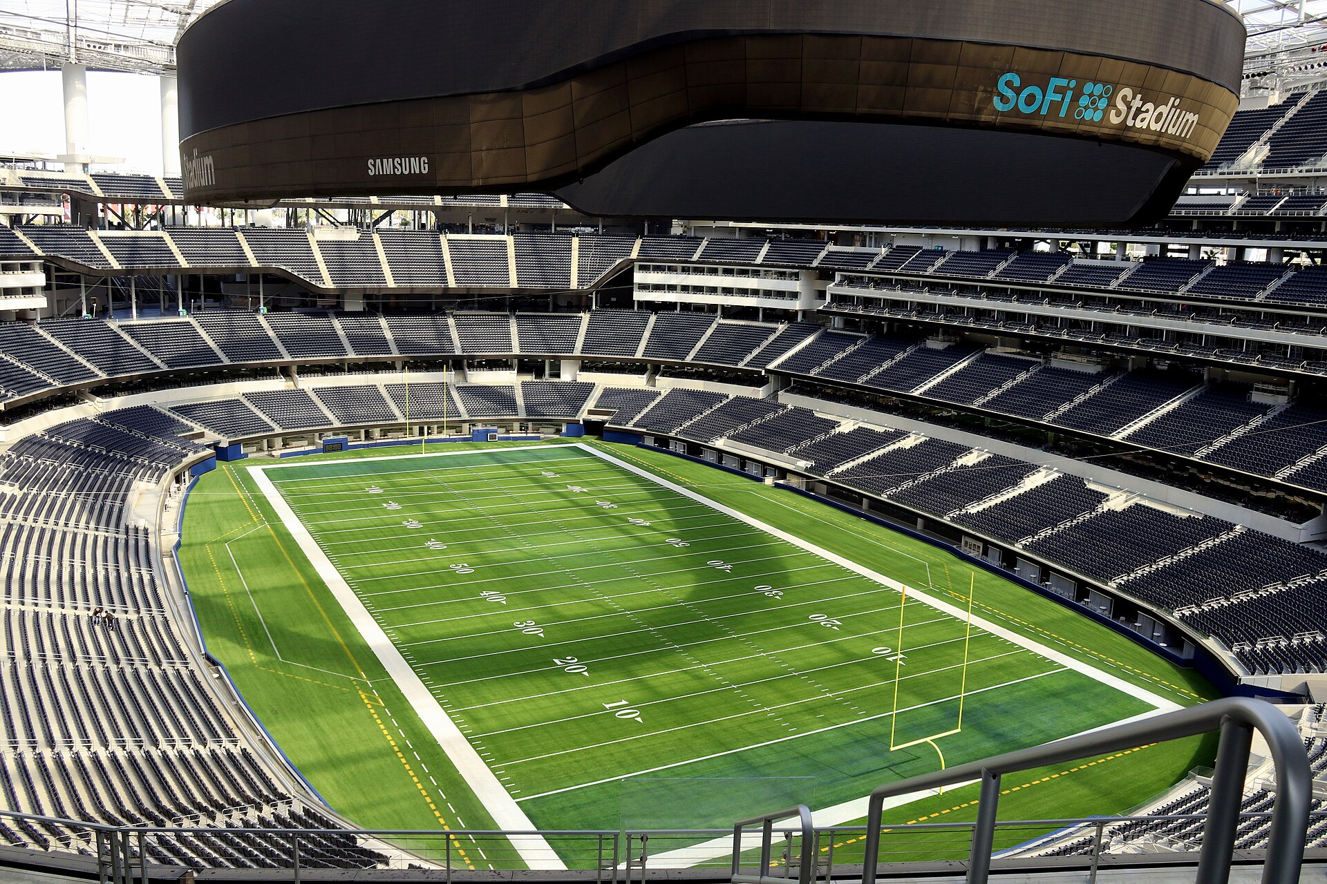An empty SoFi Stadium with its green football field, rows of dark seats and the oval video board overhead