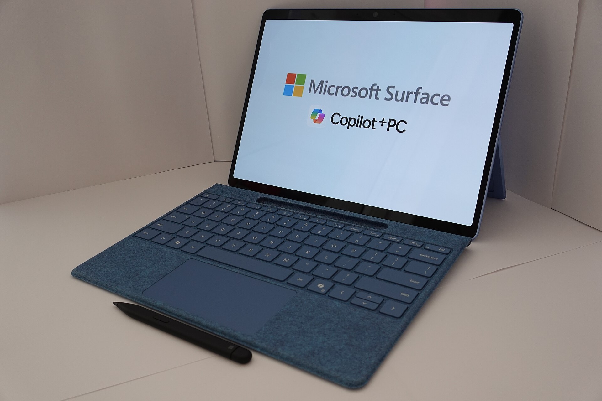 A Microsoft Surface Copilot+ PC with a blue keyboard and pen, its screen showing the Microsoft Surface logo